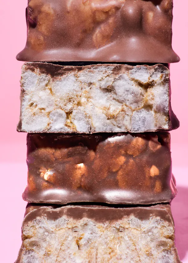 ein stapel aus vier schokoriegeln auf rosa hintergrund, wobei zwei riegel durchgeschnitten sind und eine knusprige, weiße füllung zeigen.