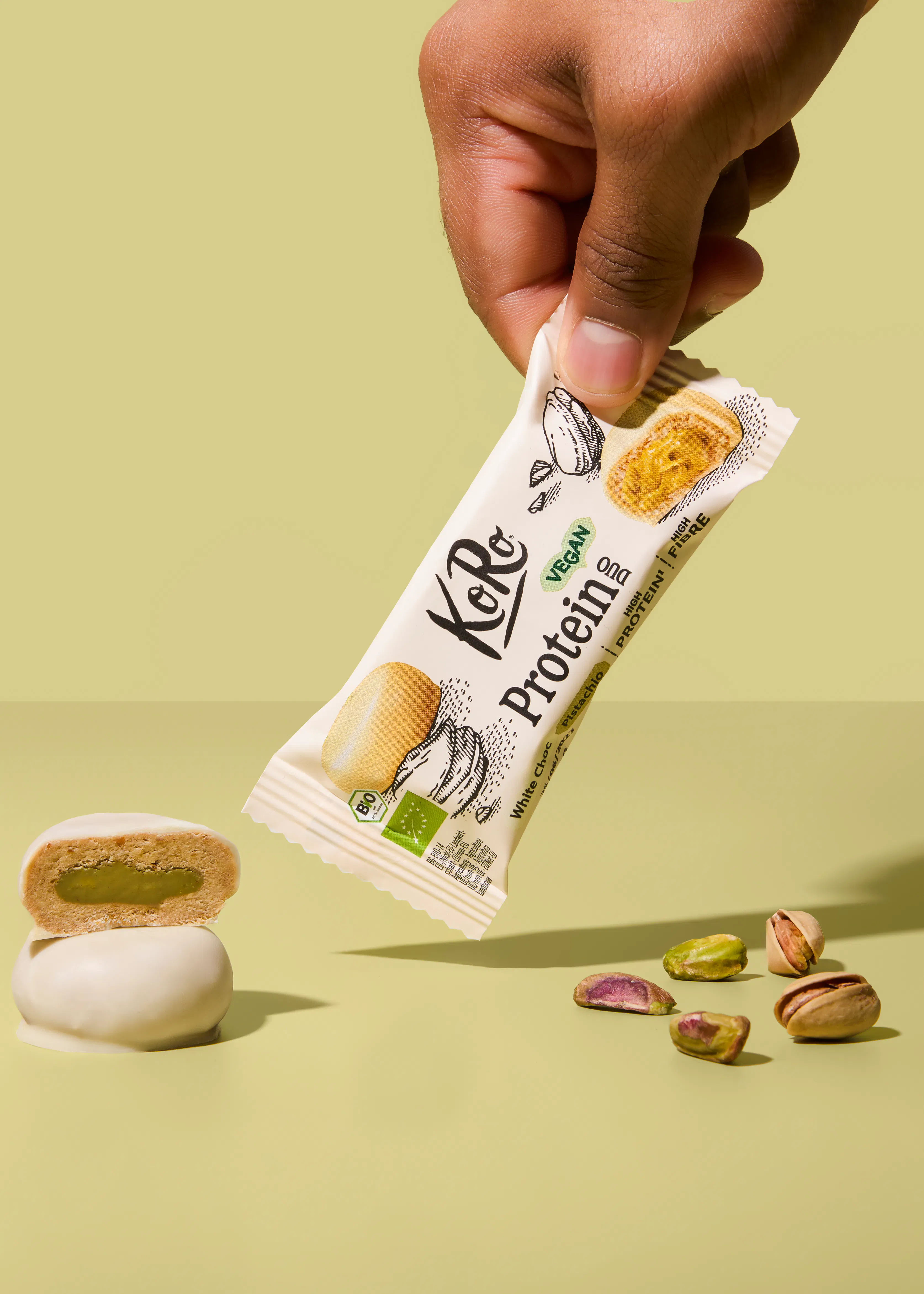 une main tient une barre protéinée koro au chocolat blanc et pistache, accompagnée d'une bouchée coupée en deux révélant une garniture verte et de pistaches éparpillées sur un fond vert clair.