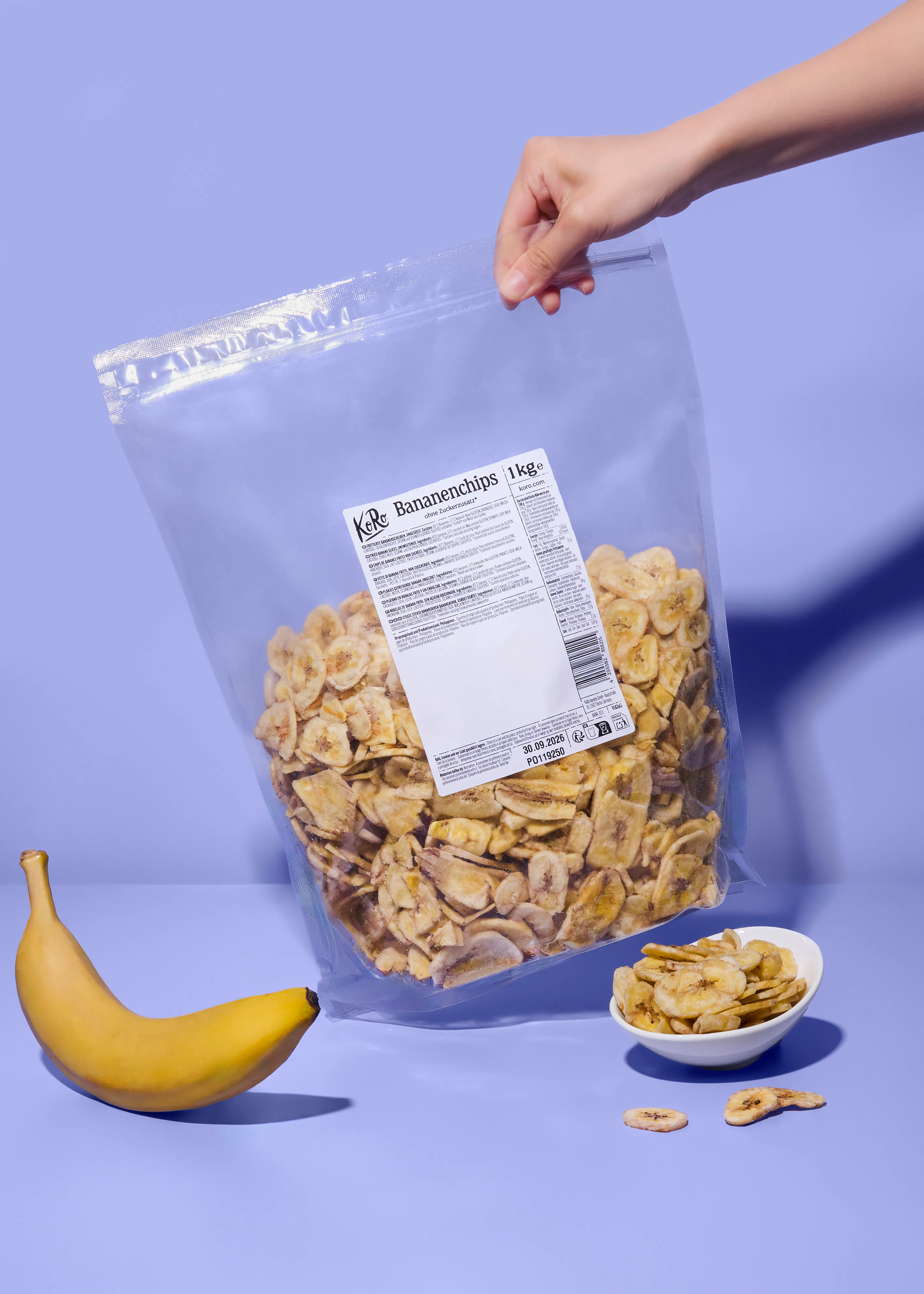 eine hand hält einen großen beutel bananenchips, daneben liegen eine frische banane und eine schale mit bananenchips.