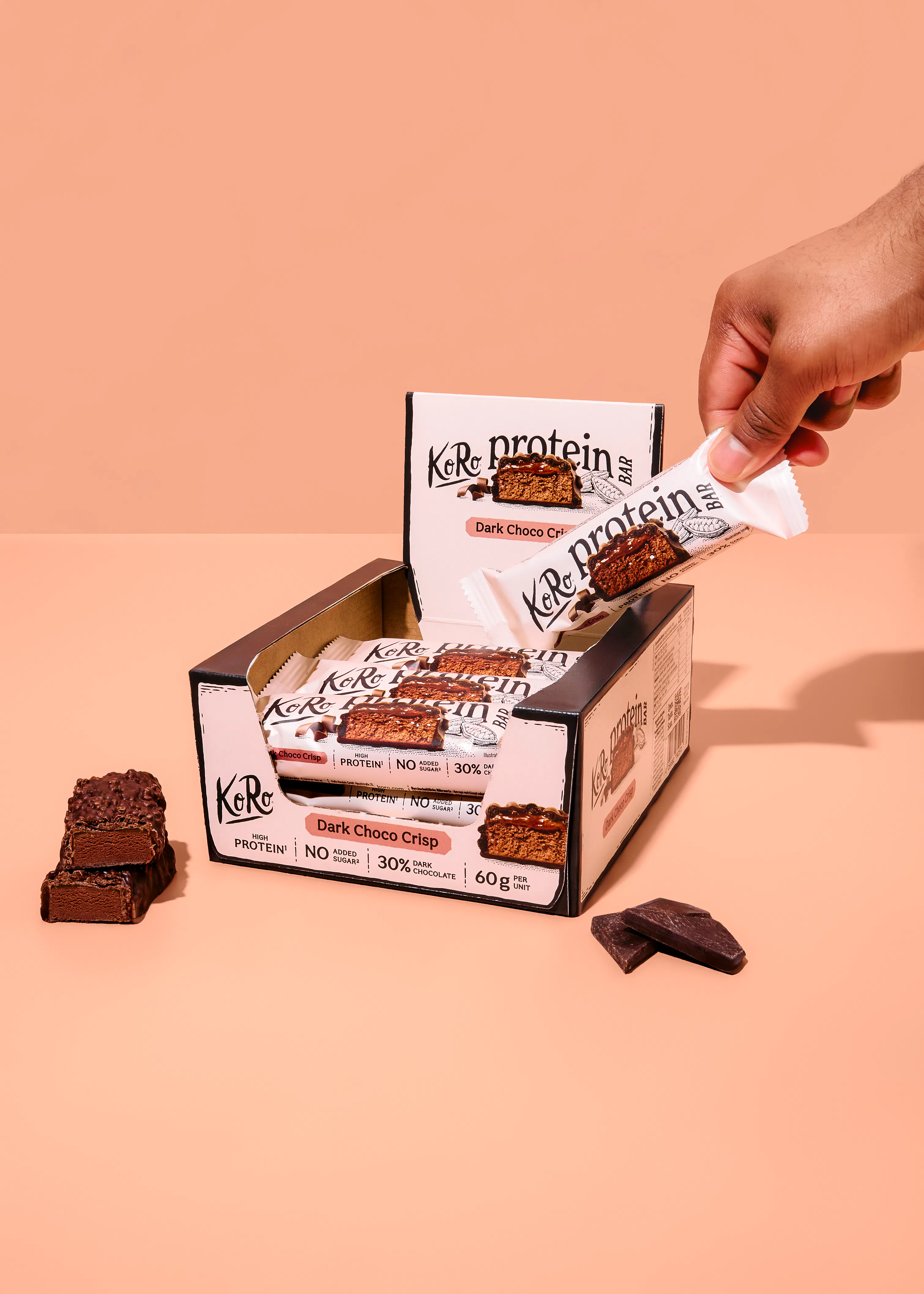 eine hand nimmt einen koro protein dark choco crisp riegel aus einer schachtel, wobei weitere riegel und schokoladenstücke sichtbar sind.
