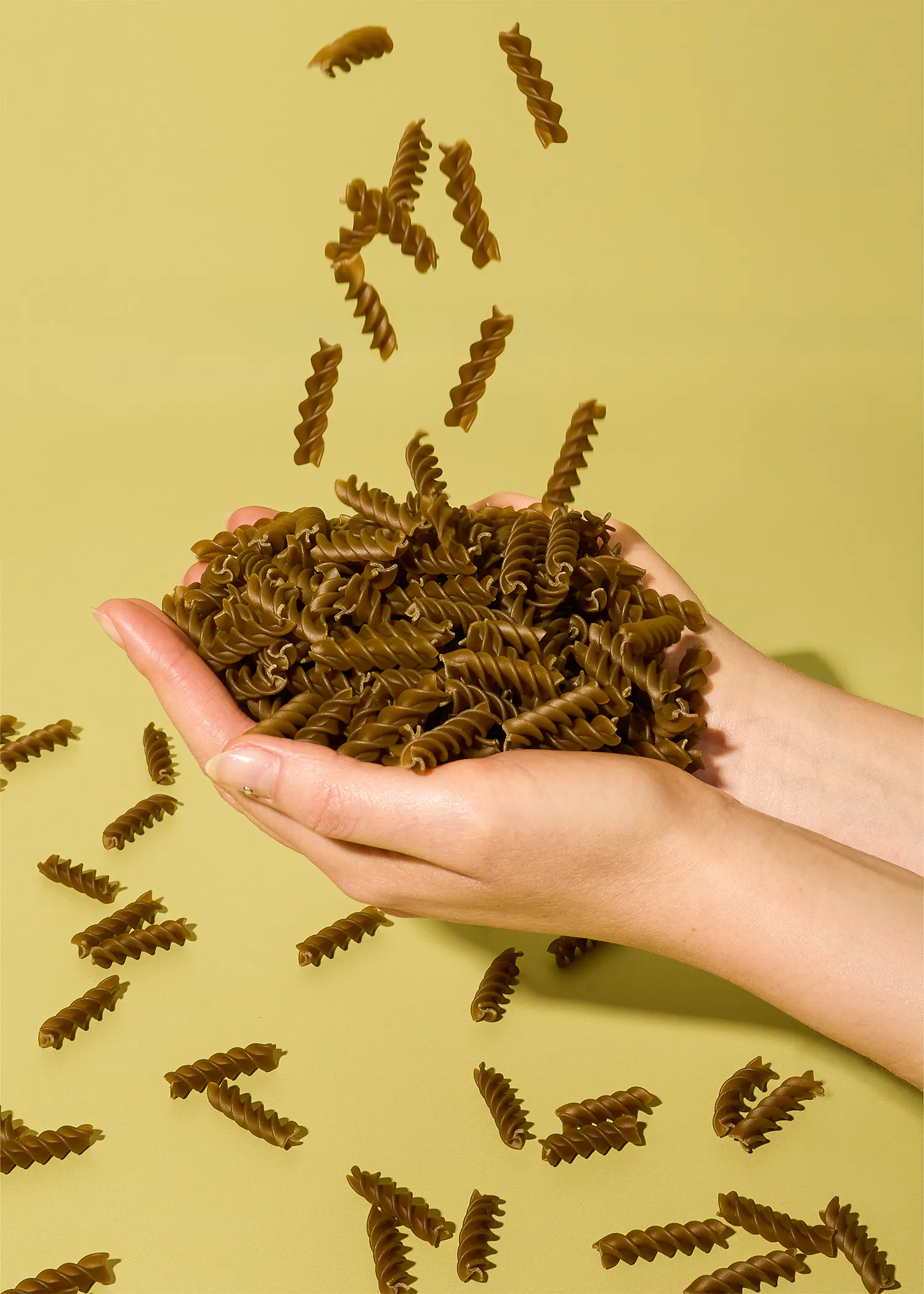 a pair of hands cupped together holding a pile of dark brown fusilli pasta with more pieces falling from above against a light green background.