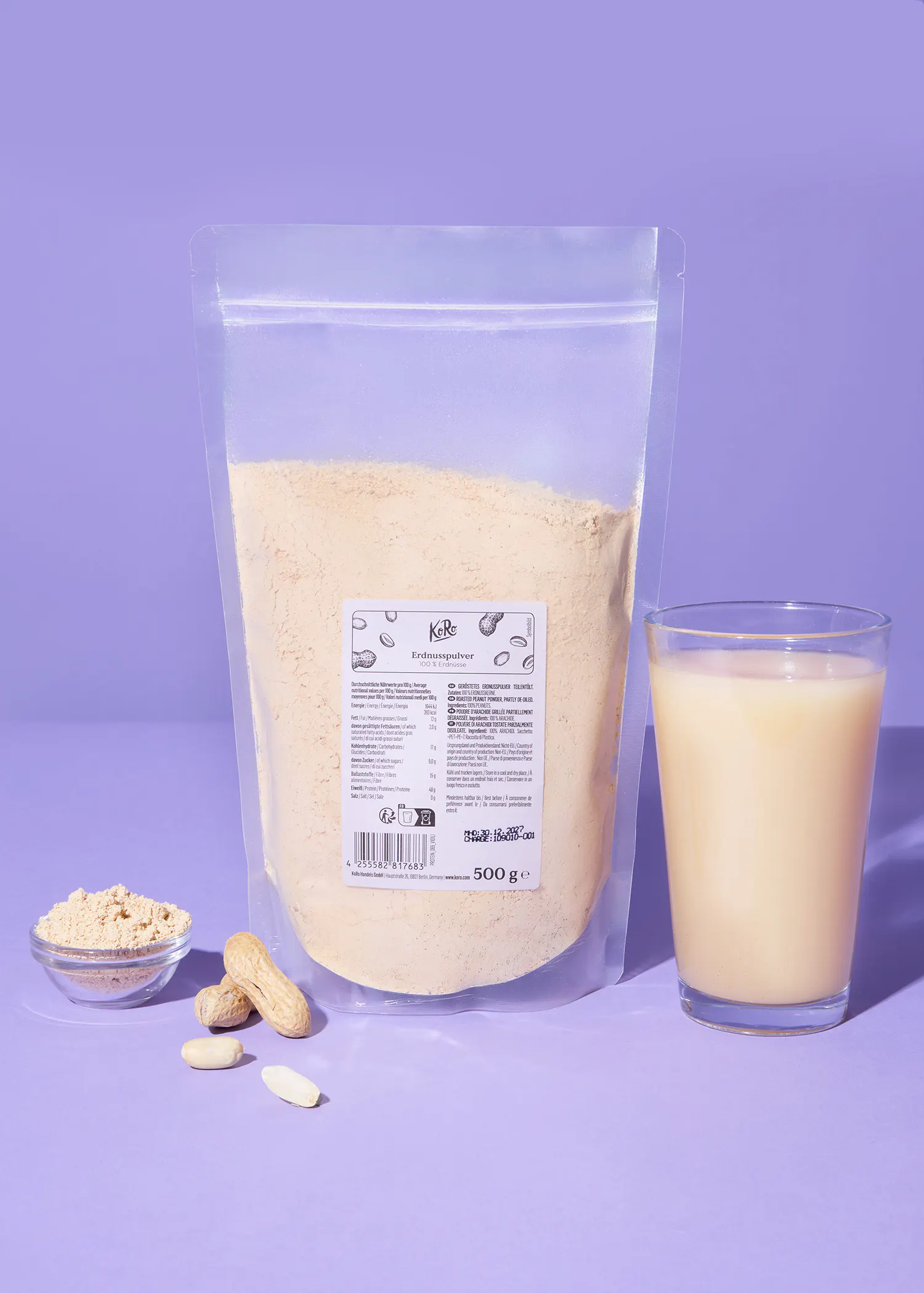 un sachet transparent de poudre d'arachide de la marque kere, un verre de boisson à base d'arachide, un petit bol de poudre et des arachides entières et décortiquées sur un fond violet.