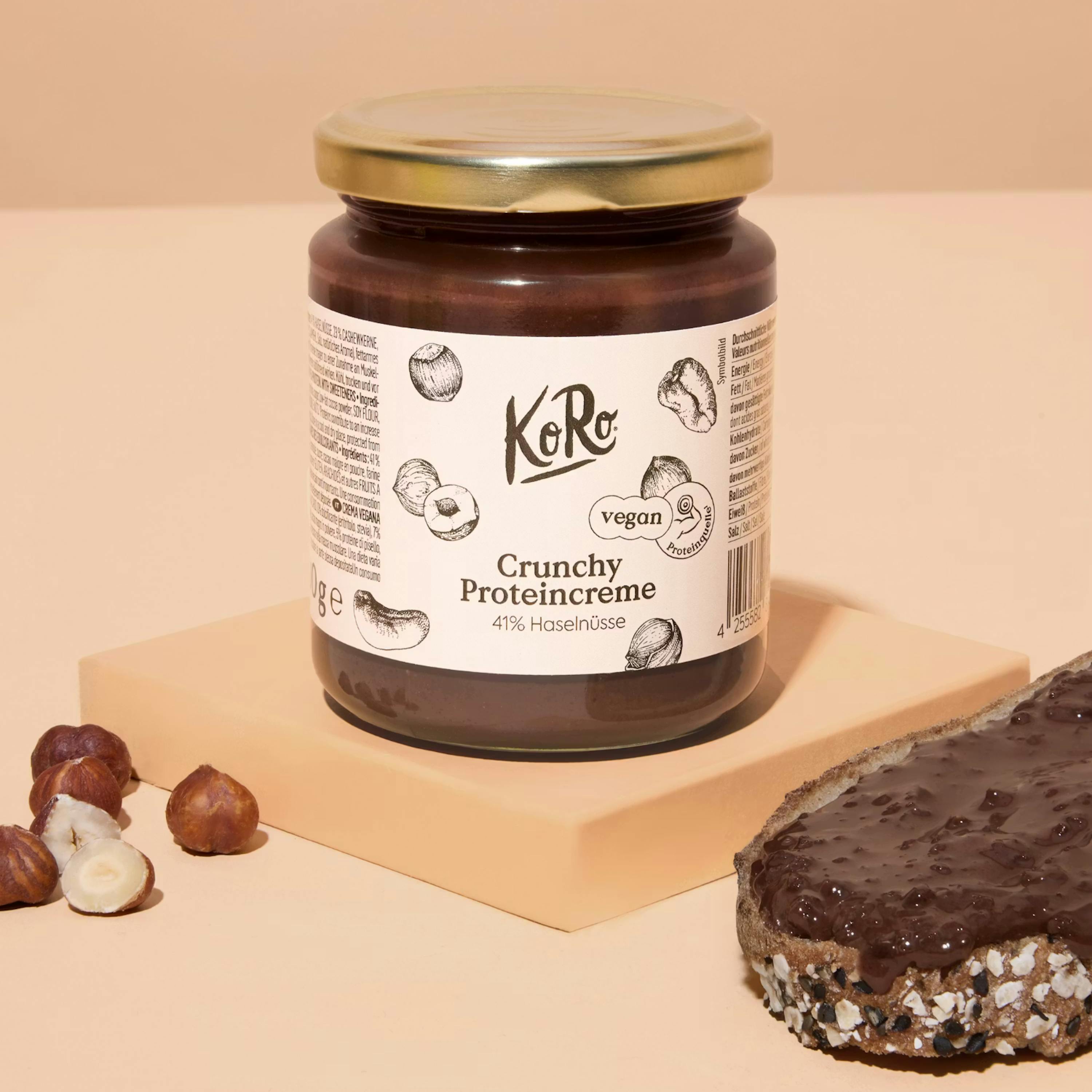 un pot de pâte à tartiner aux noisettes ko ro crunchy proteincreme végétalienne, des noisettes entières et cassées, et une tranche de pain tartinée sur une surface beige.