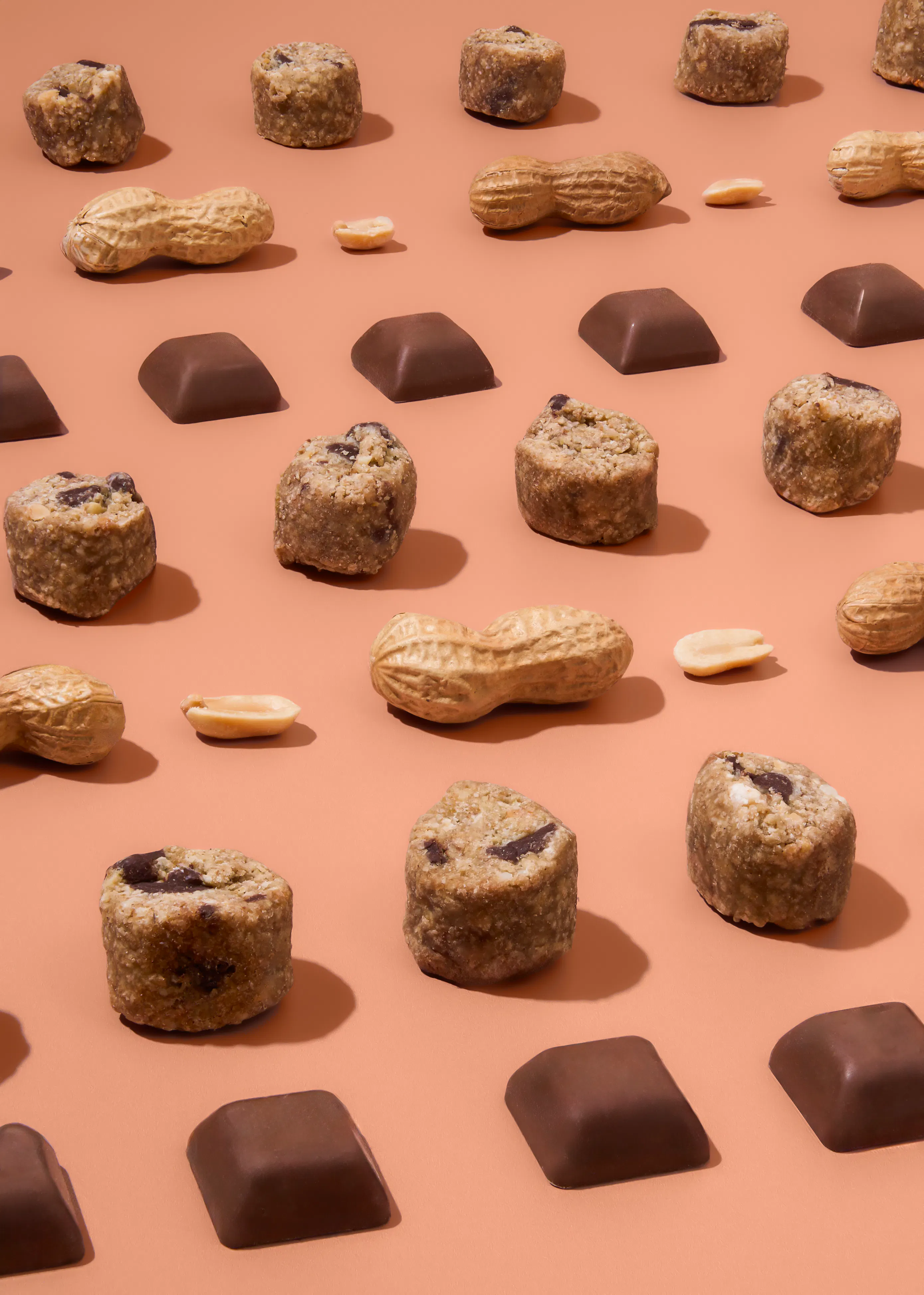 eine anordnung von erdnussbutter-bites, schokoladenquadraten und ganzen sowie geschälten erdnüssen liegt auf einem braunen hintergrund.