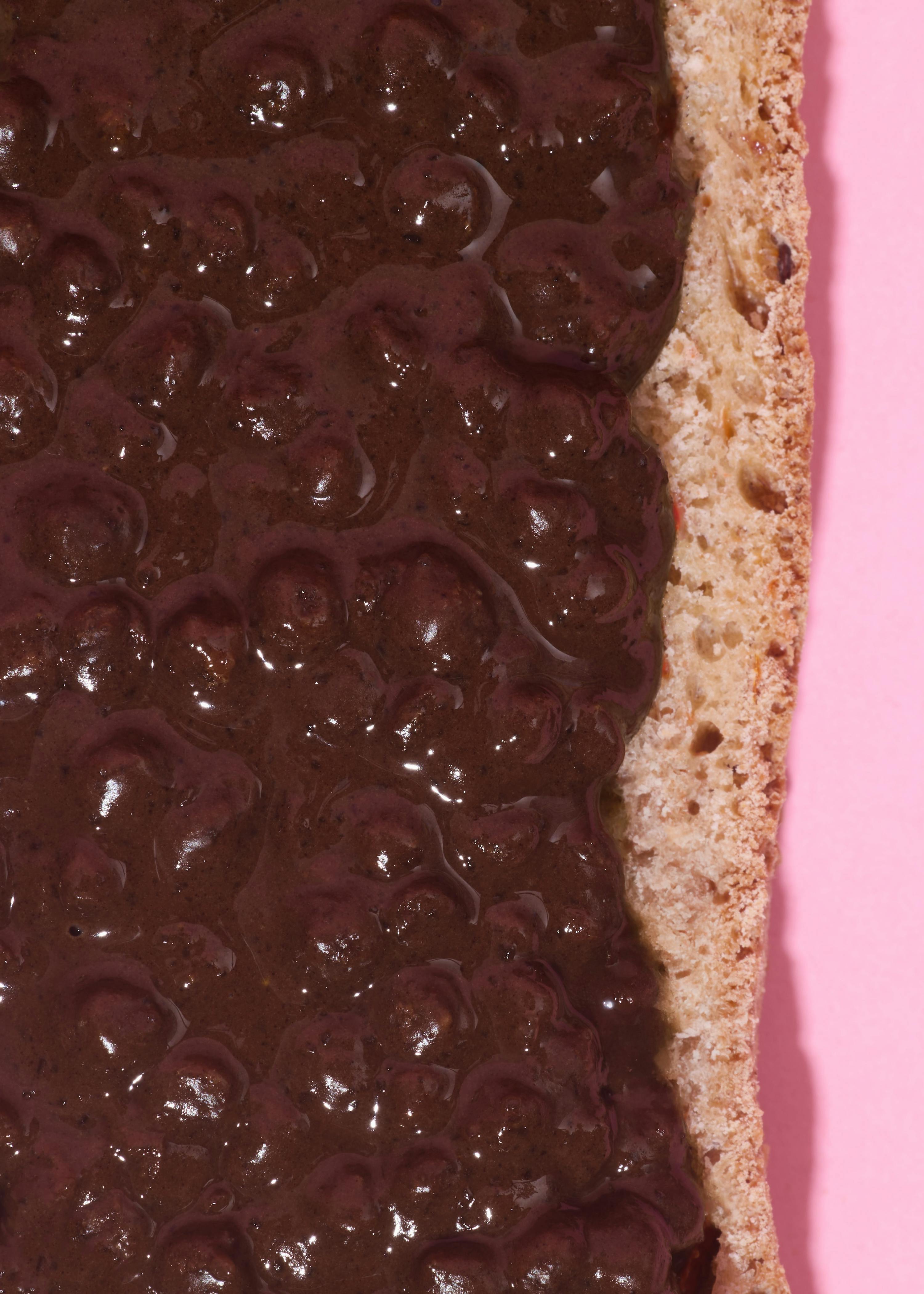 a close-up of a piece of bread covered in a thick, dark, lumpy spread, against a pink background.