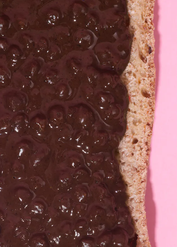 a close-up of a piece of bread covered in a thick, dark, lumpy spread, against a pink background.