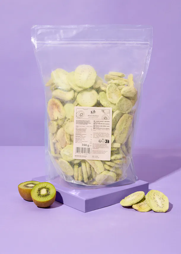 a clear plastic bag of freeze-dried kiwi slices sits on a purple surface, with two fresh kiwi halves and some loose dried slices nearby.