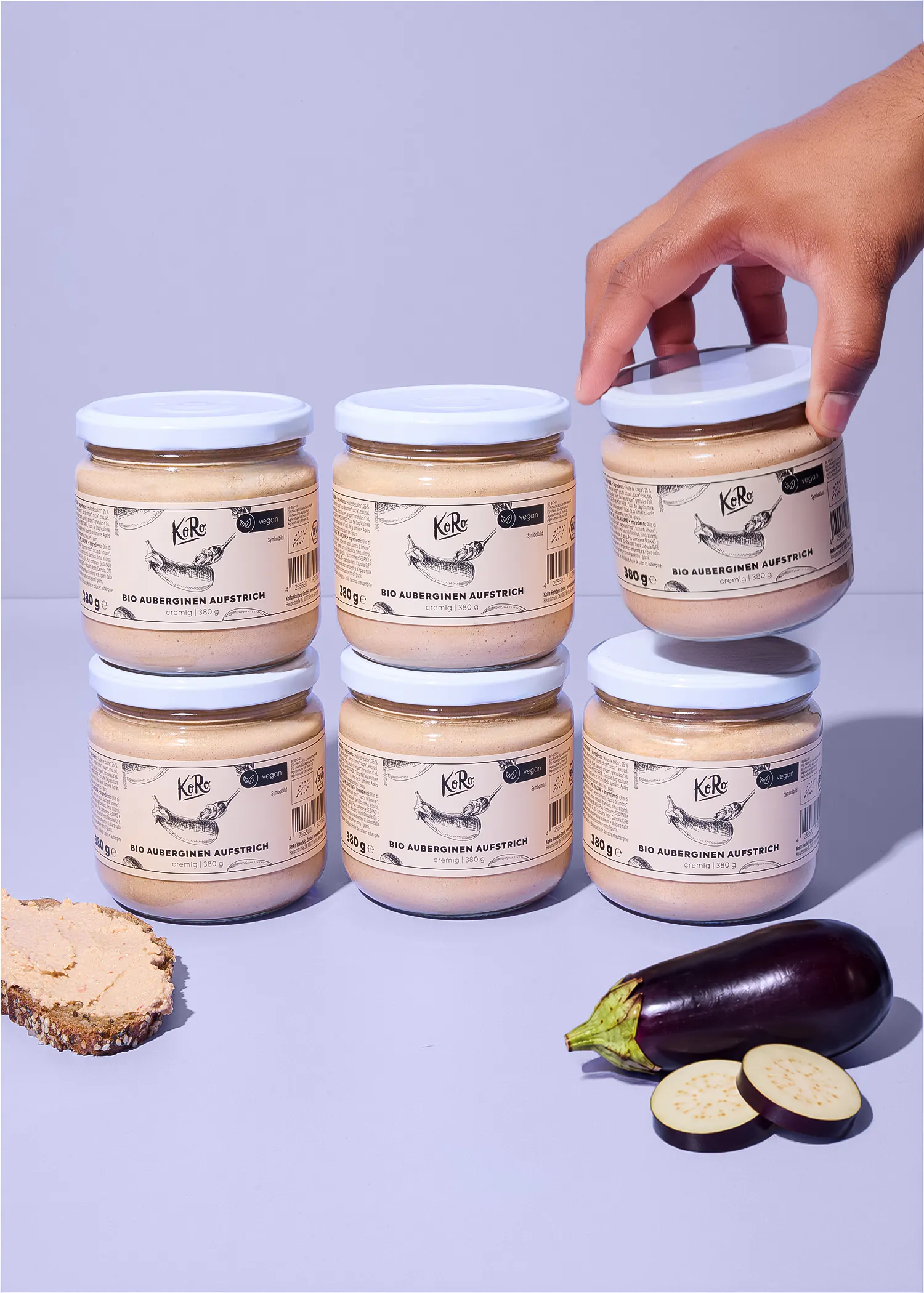 eine hand hält ein glas koro bio-auberginen-aufstrich über einen stapel weiterer gläser, daneben liegen eine ganze aubergine und zwei scheiben sowie eine mit aufstrich bestrichene brotscheibe.