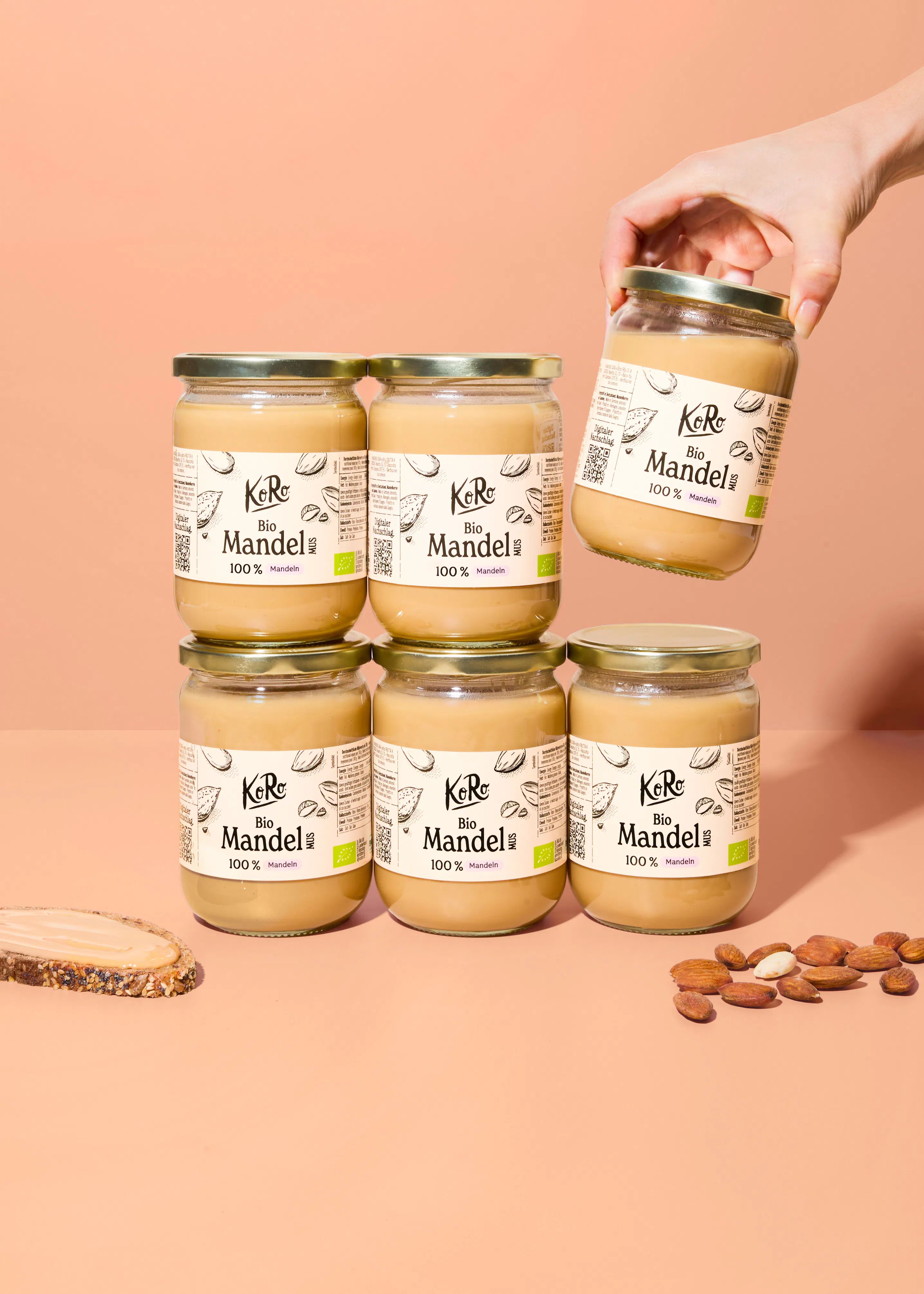 eine hand hält ein glas koro bio mandelmus über fünf weiteren gläsern, die auf einer fläche mit einer scheibe brot und einigen mandeln arrangiert sind.