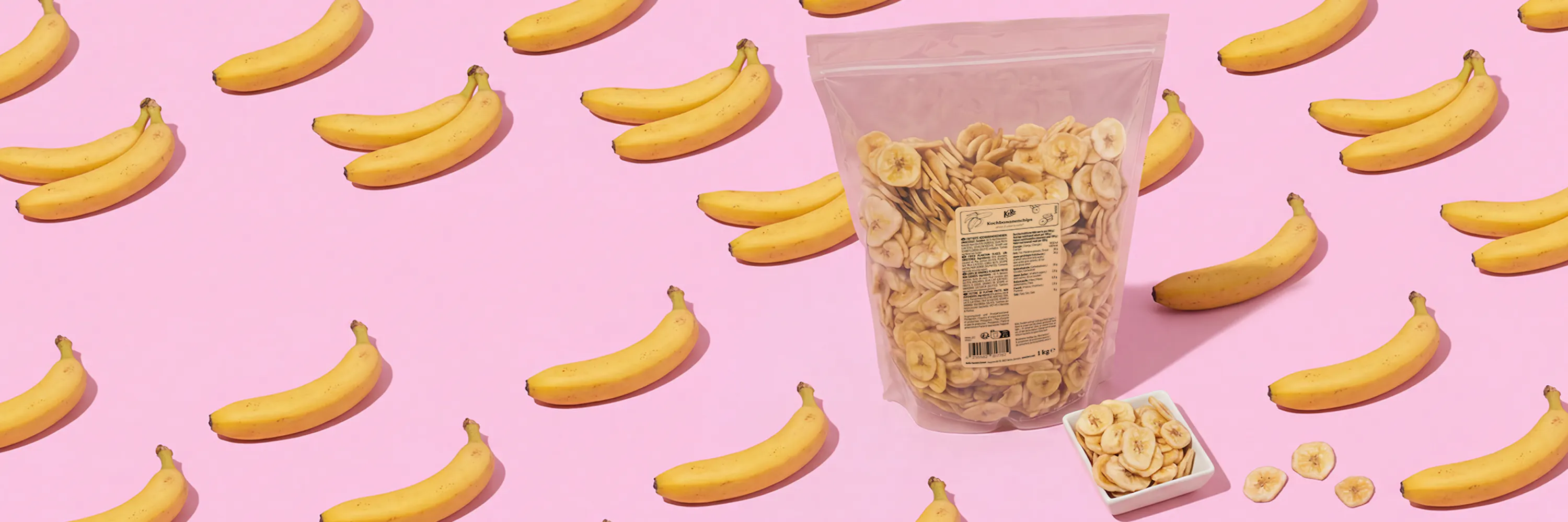 eine große tüte und eine kleine schale mit bananenchips stehen auf einem pinkfarbenen hintergrund, umgeben von frischen bananen.