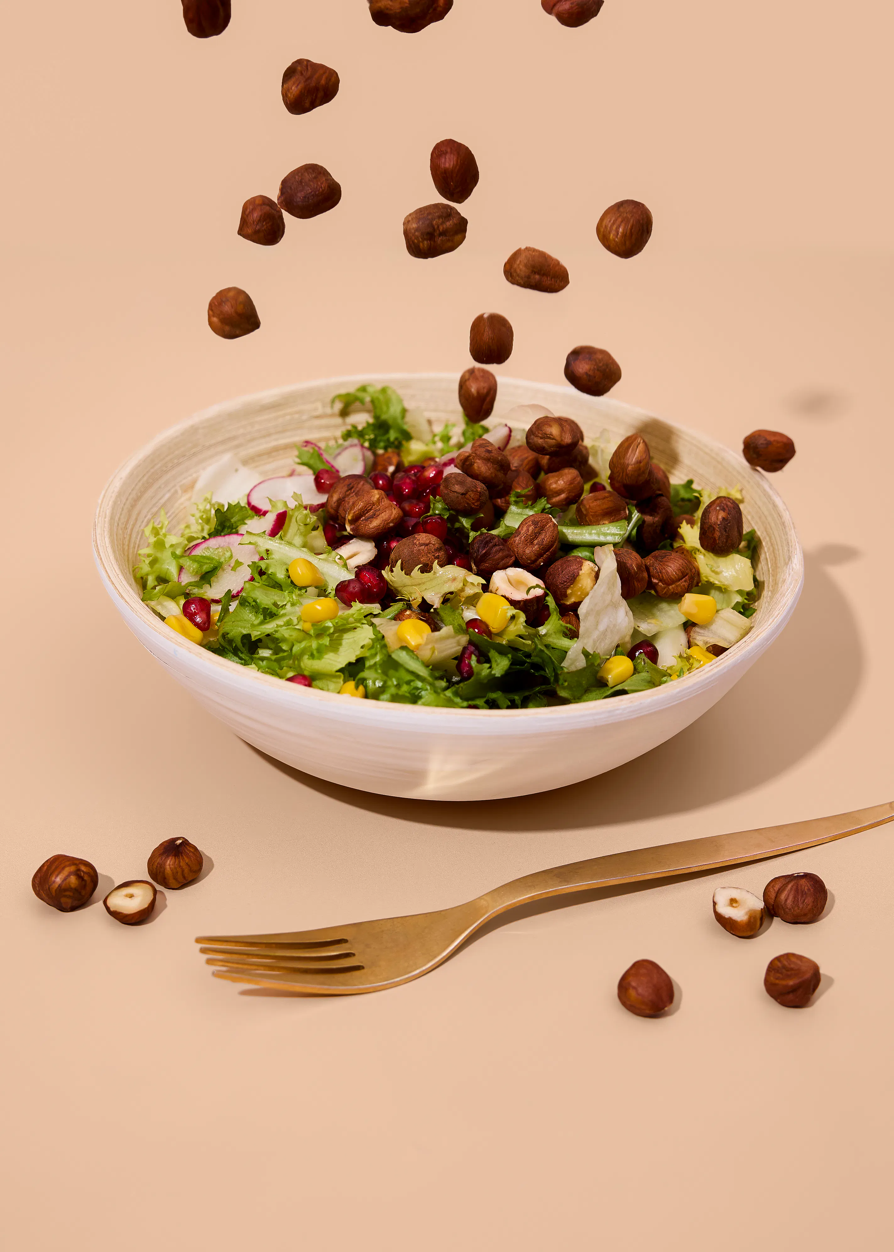 ein salat in einer schüssel mit salat, mais, granatapfelkernen und radieschen, wobei haselnüsse in den salat fallen und einige neben einer goldenen gabel auf einem beigefarbenen tisch liegen.