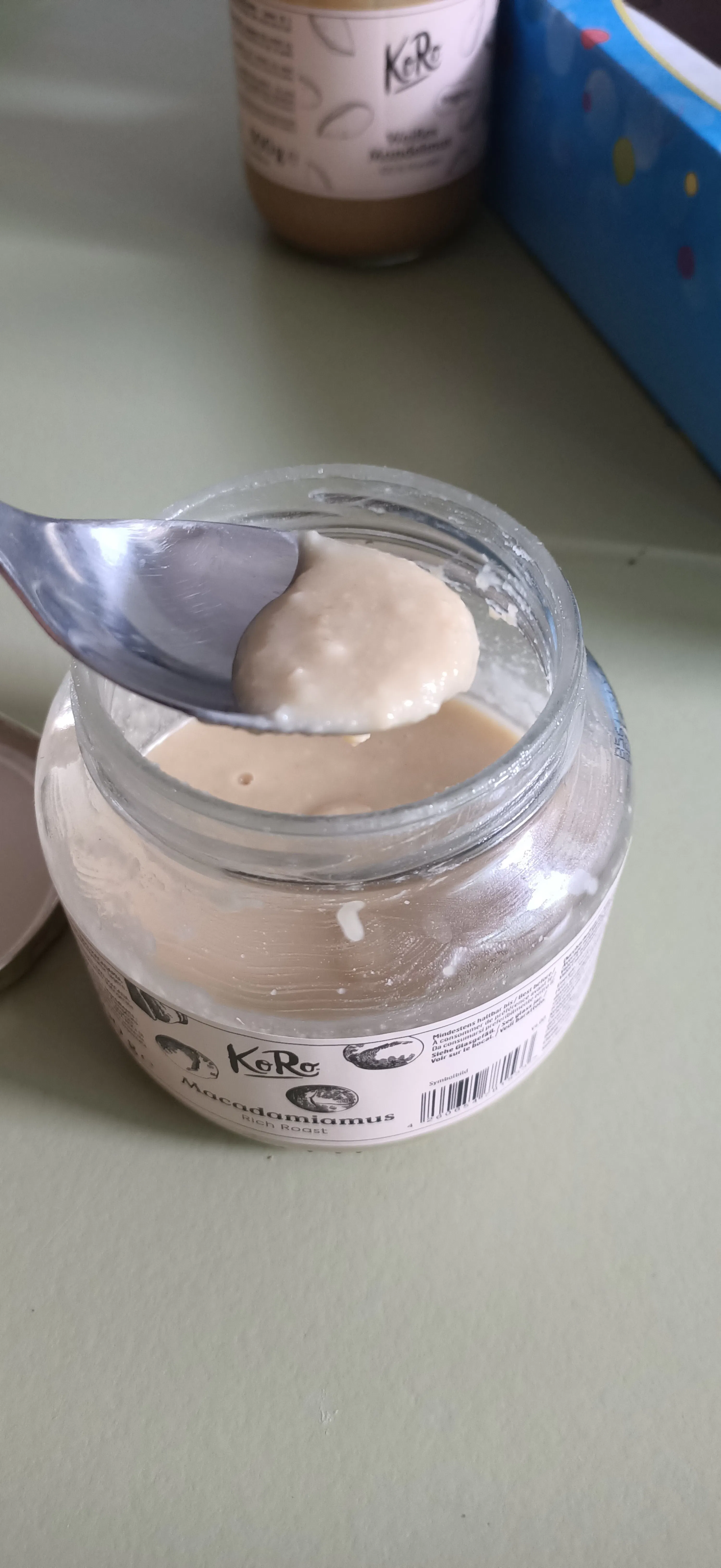 ein löffel mit hellem, cremigem macadamiamus wird aus einem glas mit der aufschrift „koro macadamiamus rich roast“ gehoben.